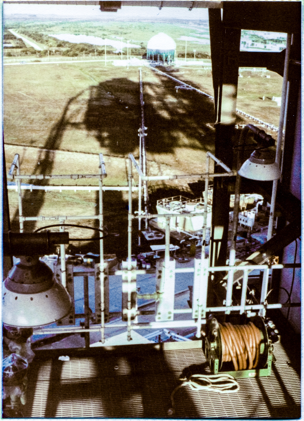 Image 034. Viewed 120 feet above the distant wilderness from the lower reaches of the Rotating Service Structure, the combined shadows of the two towers, the RSS and the FSS, at Space Shuttle Launch Complex 39-B, Kennedy Space Center, Florida, span a full quarter mile, all the way out to the Liquid Oxygen Dewar at the Pad Perimeter. In the near foreground, one of the numerous emergency-shower/eyewash stations is visible, which is equipment for personnel decontamination via copious flushing with water in the event of a Hypergol spill. Photo by James MacLaren.